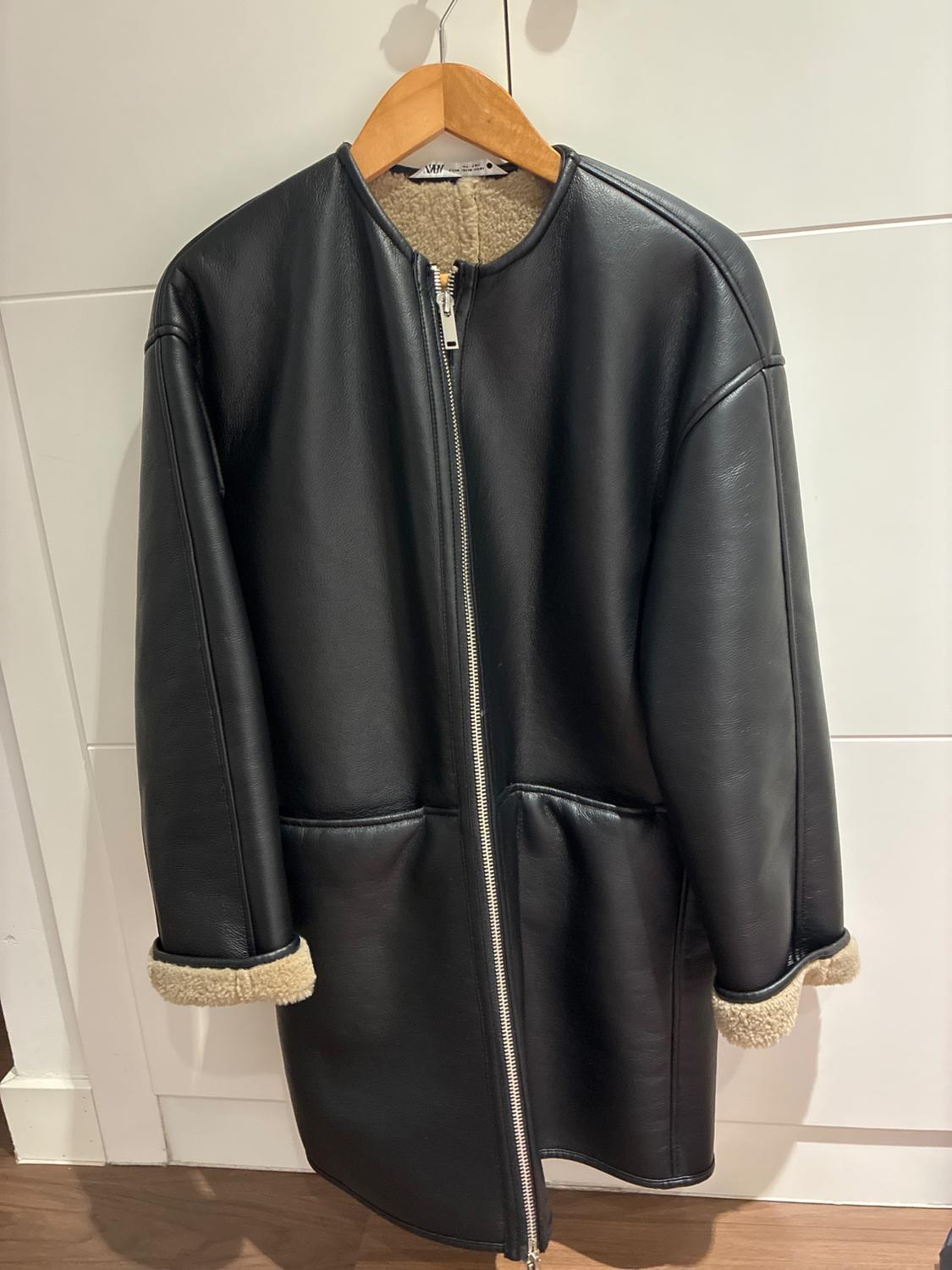 Black collarless zip-up coat with light-coloured lining on the cuffs, hanging on a wooden hanger against a white door. Text: ZARA.