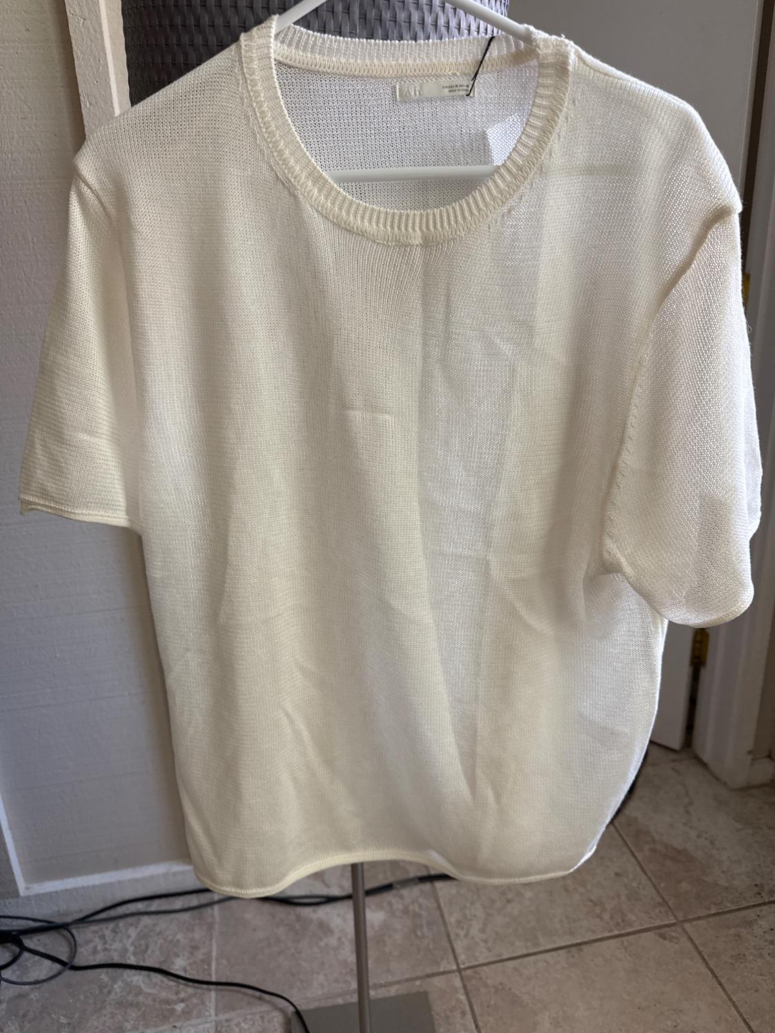 Short-sleeved, round-neck knitted T-shirt in Ecru, hanging on a white hanger in a domestic setting.