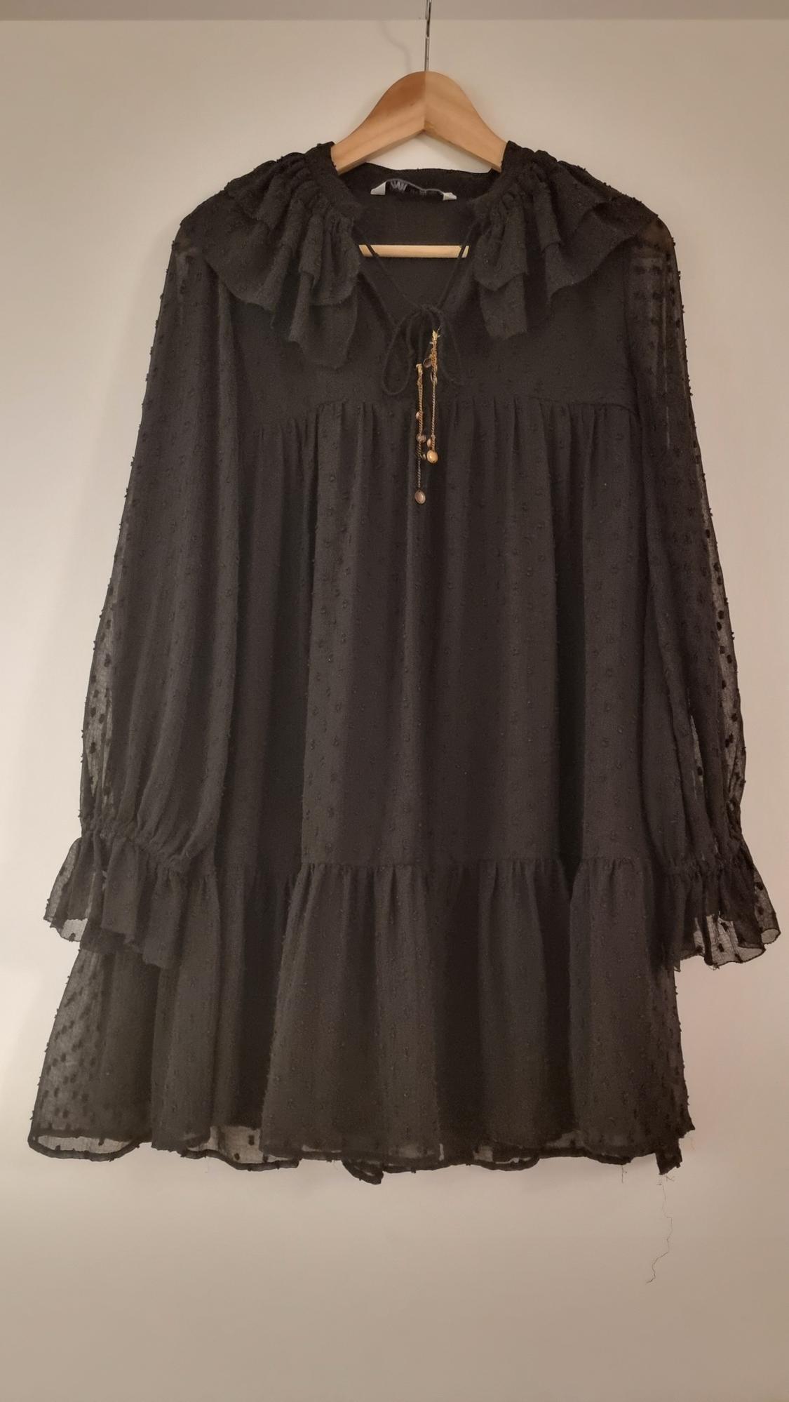 Black dress with polka dot texture, long sleeves and frills on the collar, hanging on a wooden hanger against a white background.
