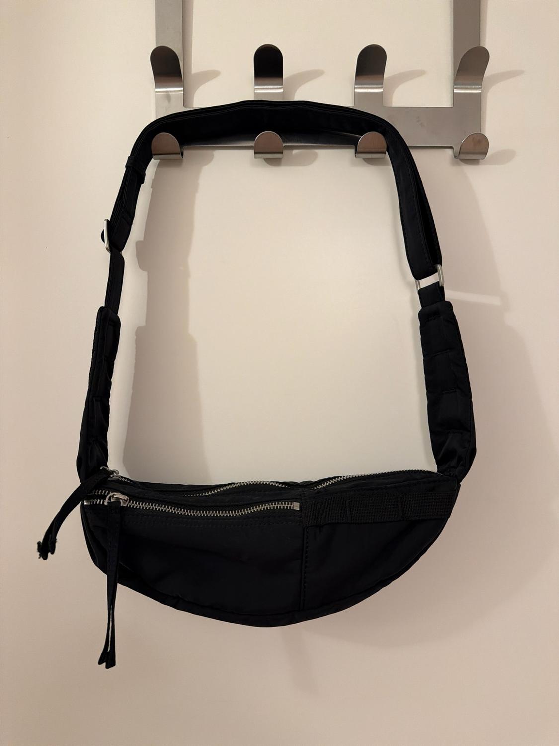 Black handbag hanging on a metal coat rack against a white wall. Curved, cross-body style design with a wide strap and front zips.