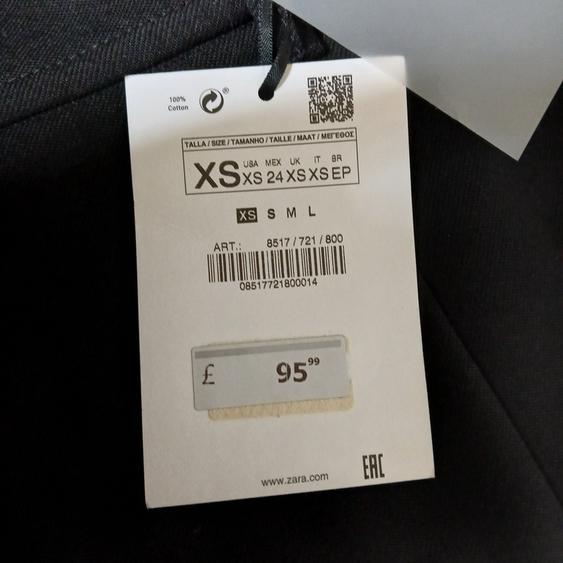 Clothing label on dark fabric, shows size XS, price £95.99, 100% cotton, barcode and website www.zara.com.