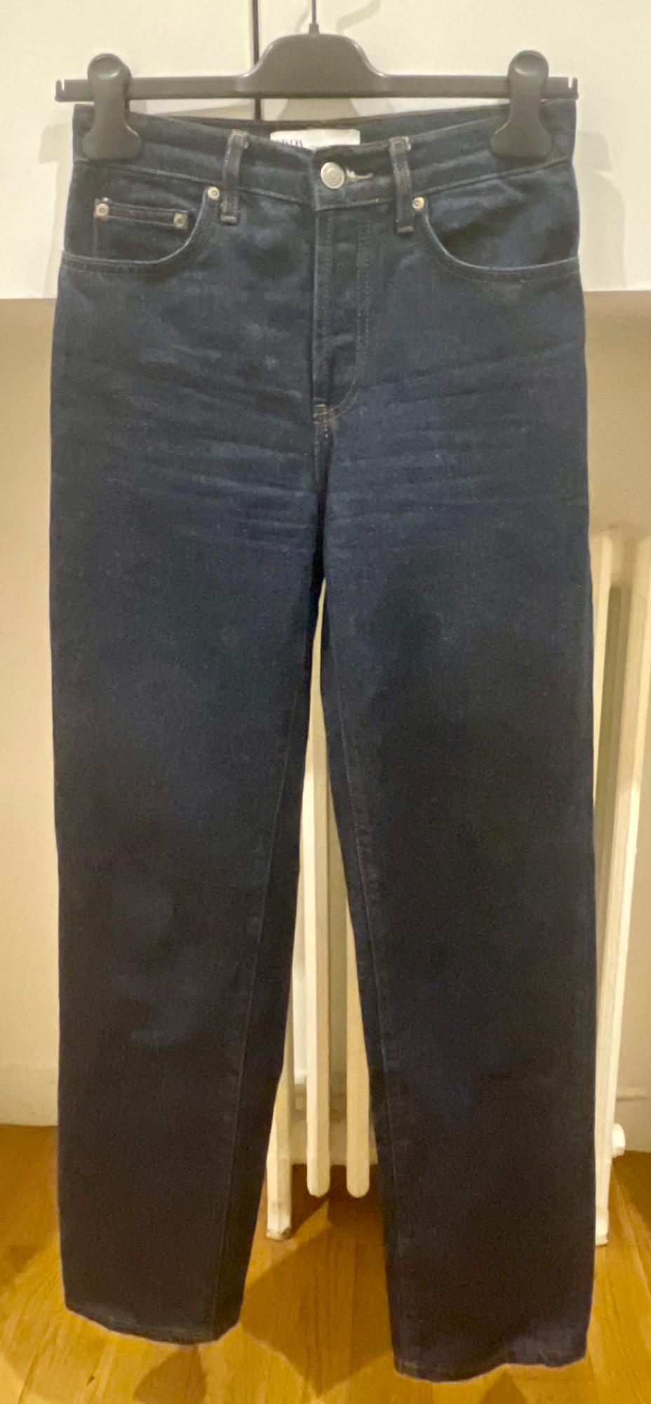 Dark blue jeans hanging on a black hanger against a white background and wooden floor.