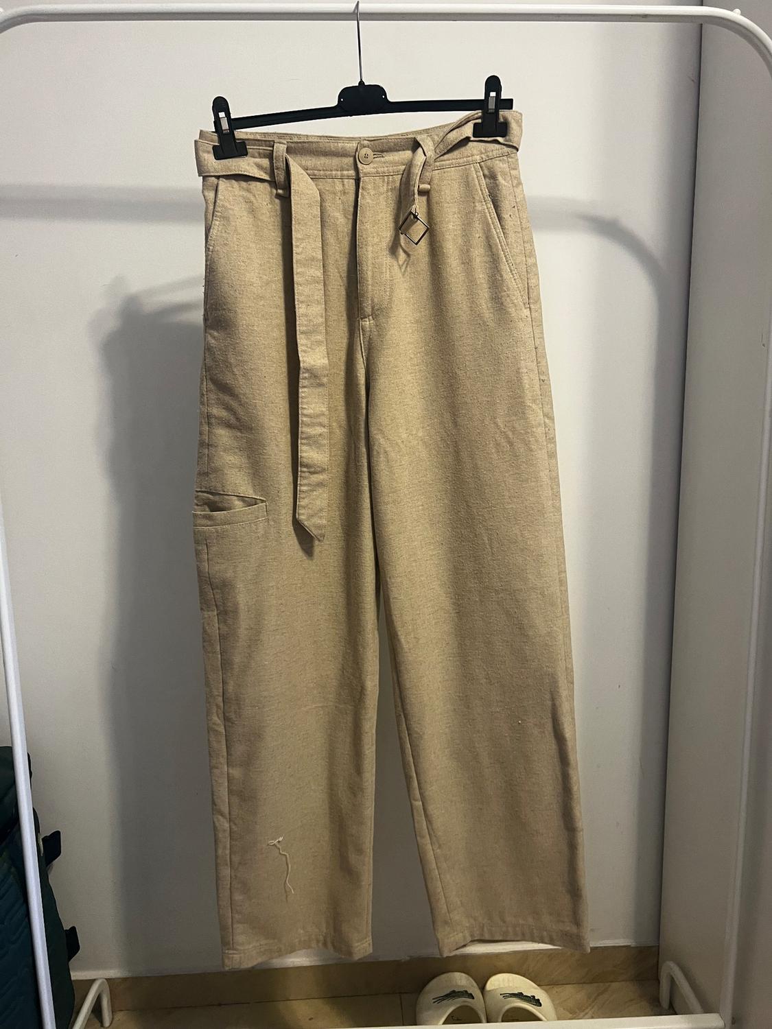 Beige trousers hung on a black hanger, with a matching belt. White background and light wooden floor.