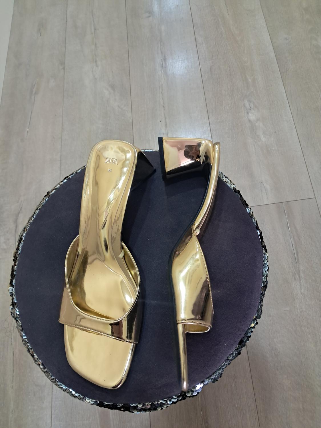 Gold high-heeled sandals on a black sequined stool. Light wooden floor visible in the background.