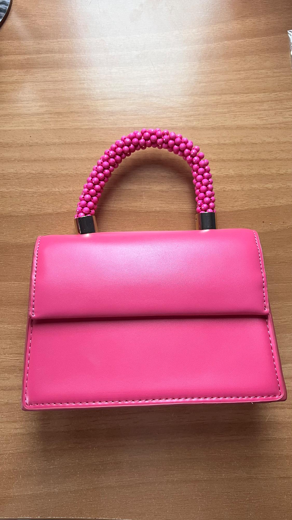Pink handbag on a wooden table, with a pink beaded handle and gold metallic details. Structured and modern design.