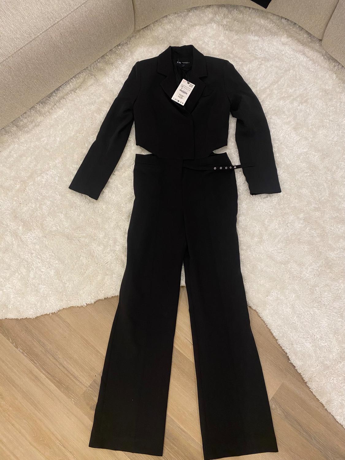 Two-piece black suit with a cropped jacket and wide-leg pants, on a white rug and hardwood floor. Visible tag.
