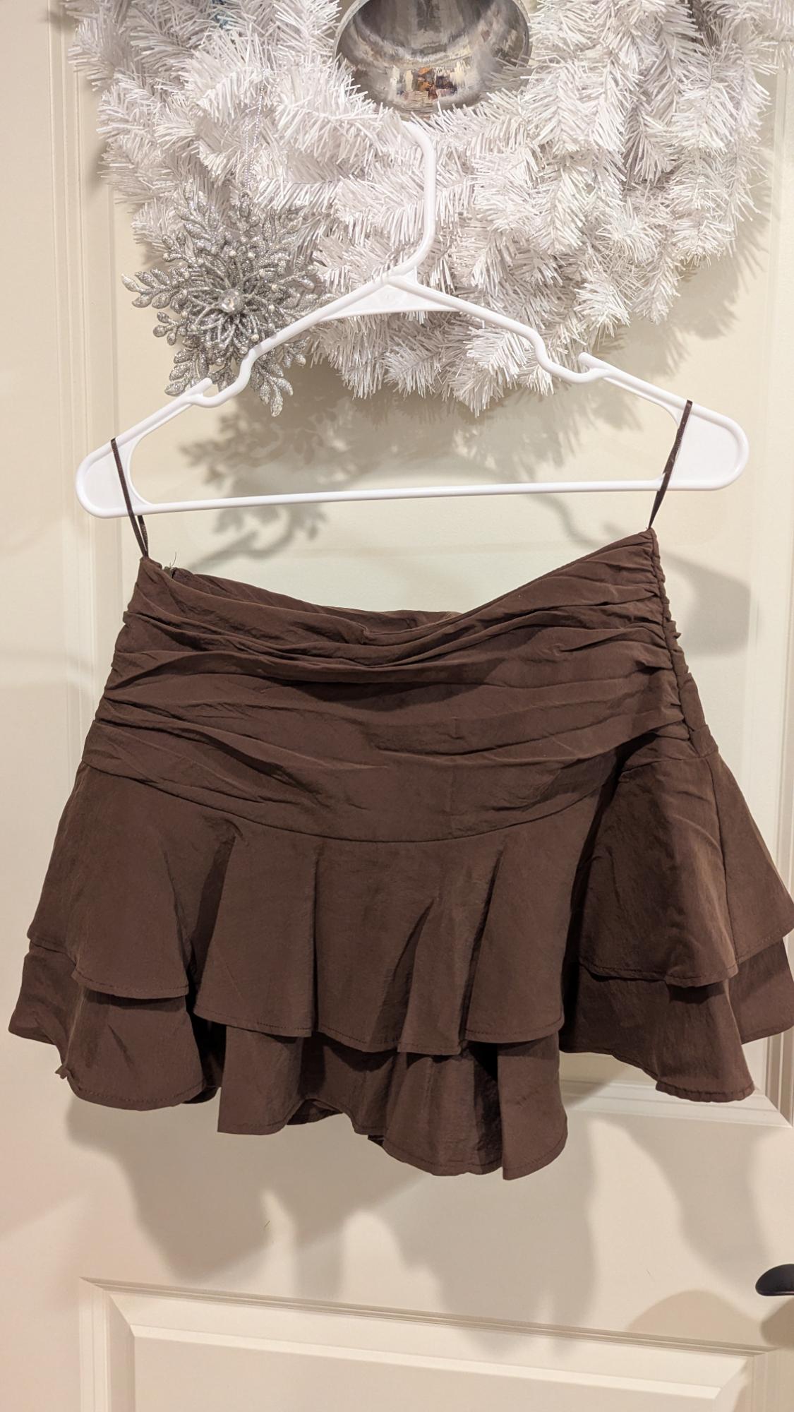 Brown ruffled skirt hanging on a white hanger, in front of a white decorative wreath with silver accents.