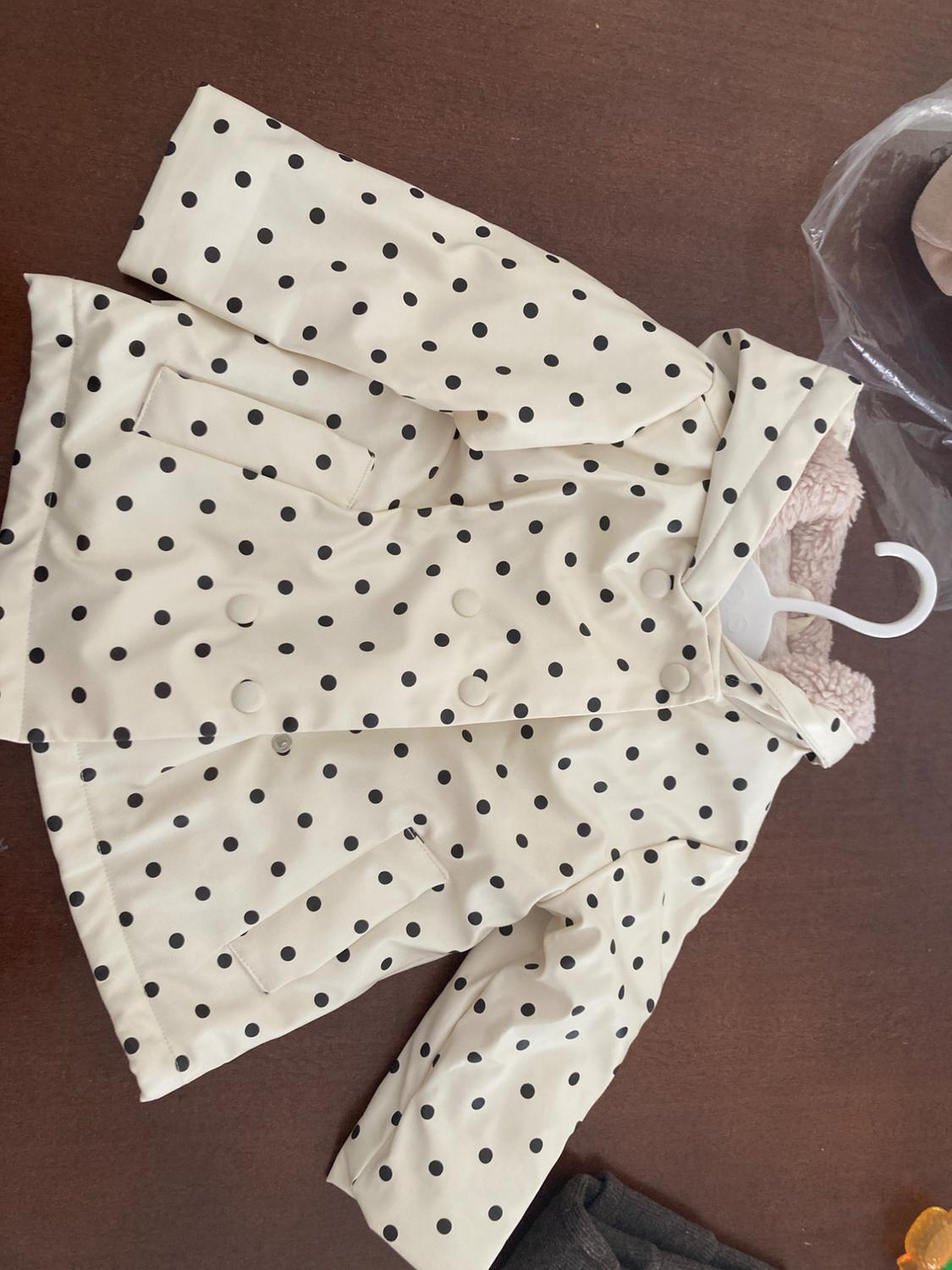 Children's white coat with black polka dots and a hood, hanging on a hanger on a brown surface.
