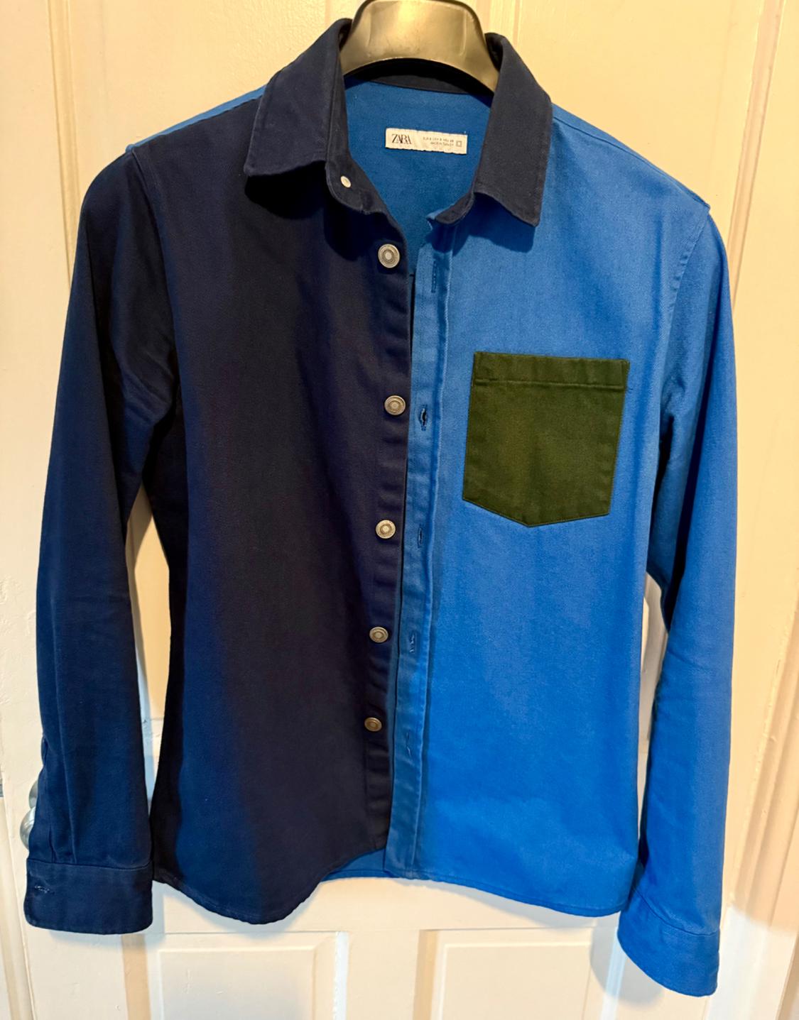 Long-sleeved shirt with a two-tone design in dark blue and light blue, with a green pocket on the chest, hanging on a hanger.