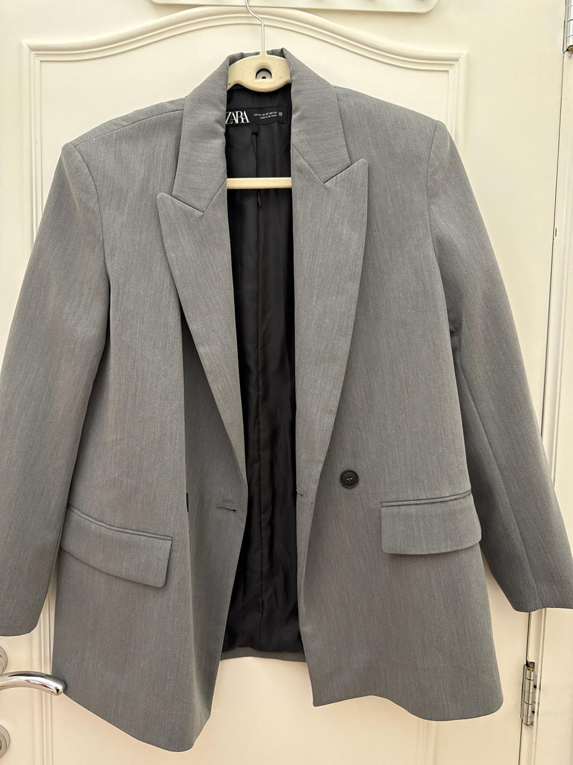 Grey blazer hanging on a hanger over a white door. Its black lining, flap pockets and the label with the text ZARA stand out.