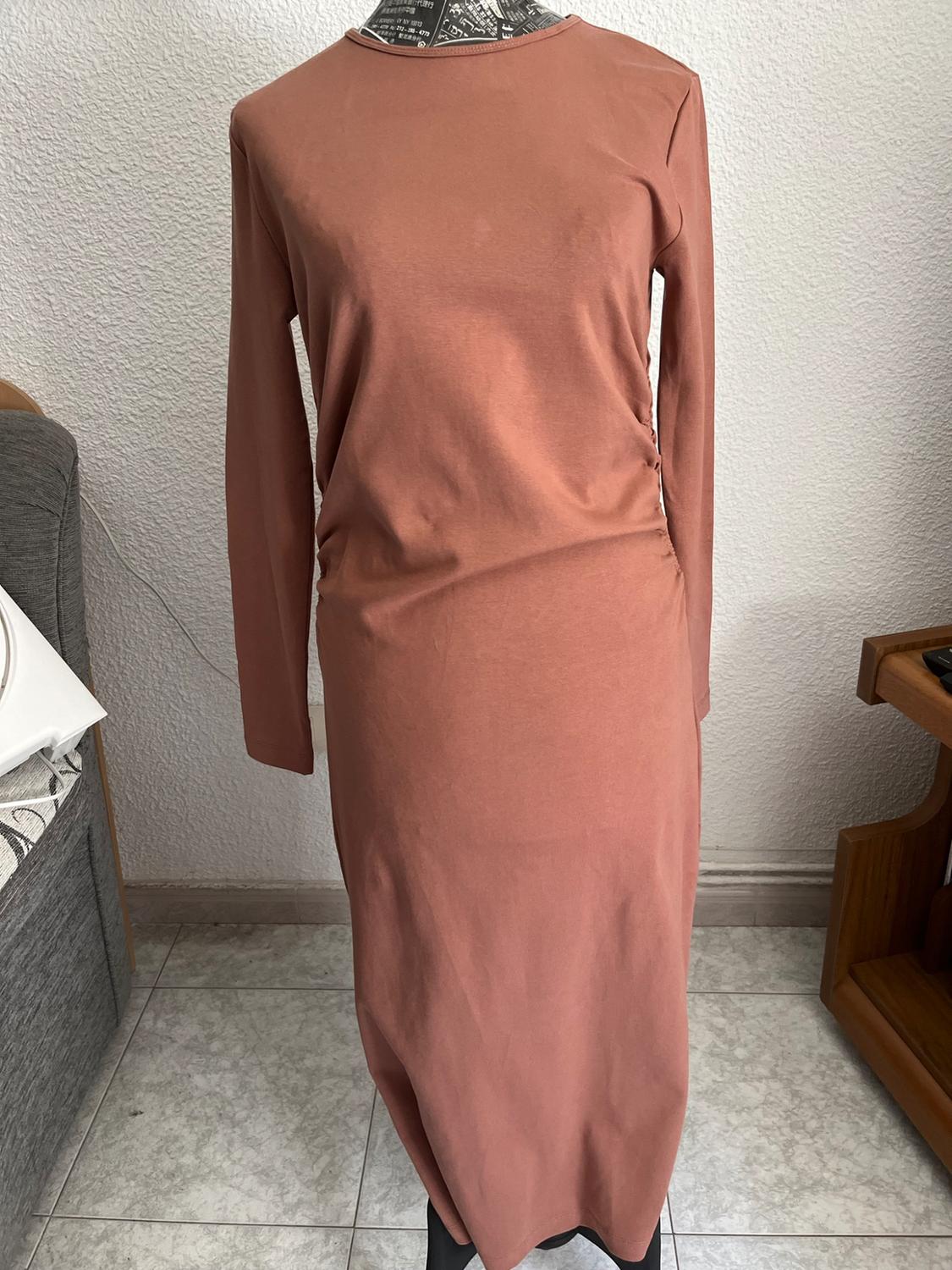 Mannequin in a casual interior wearing a long-sleeved, terracotta maxi dress with a round neck and side ruching, next to a grey sofa.