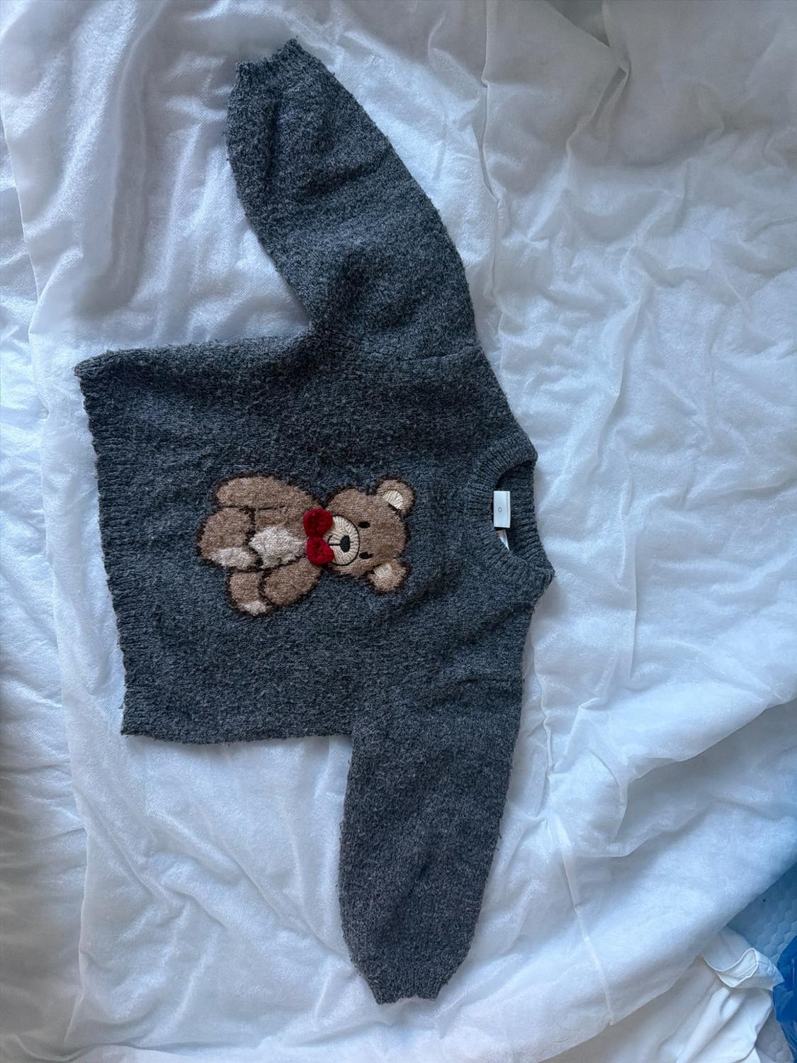 Grey knitted jumper with a teddy bear and red bow, on a crumpled white sheet.