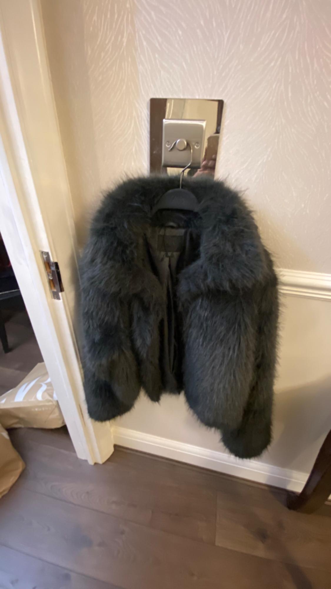 Black faux leather jacket hanging on a hanger against a light textured wall, next to a wooden door and floor.