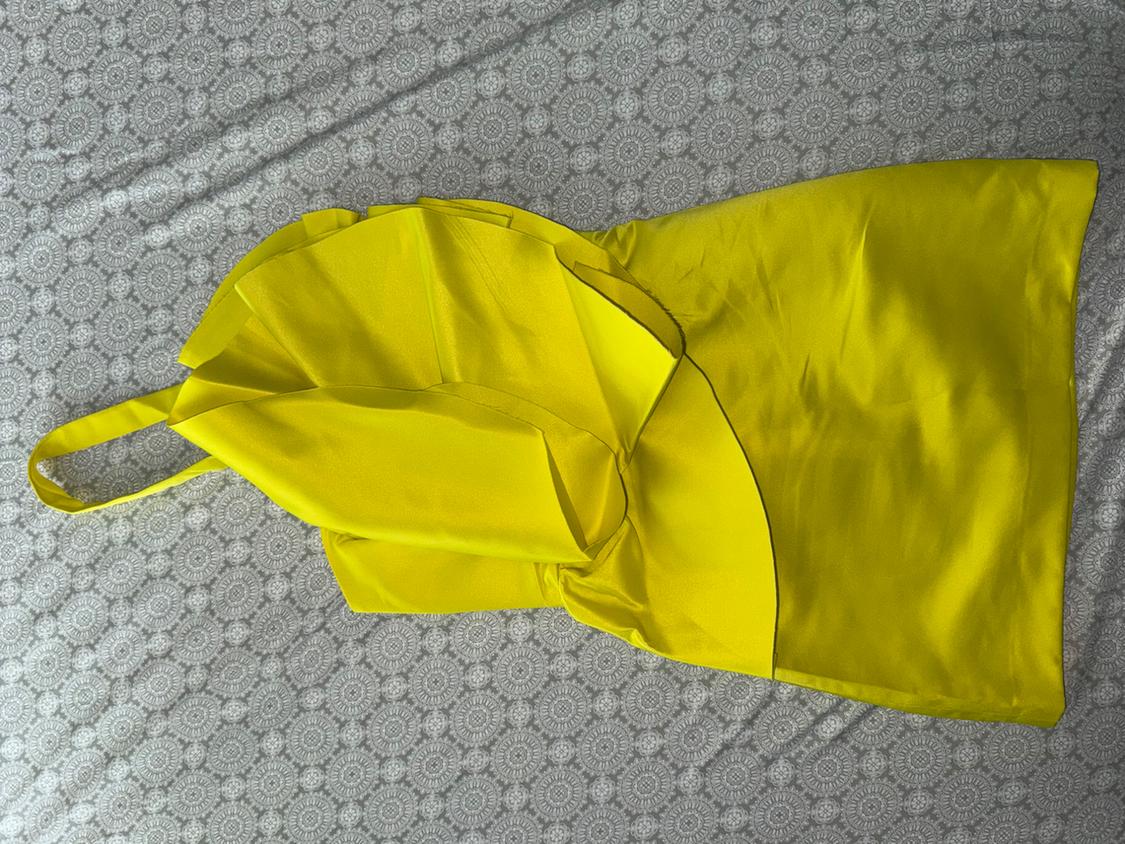 Vibrant yellow dress with an asymmetrical design and a single strap, placed on gray fabric with a repetitive circular pattern.