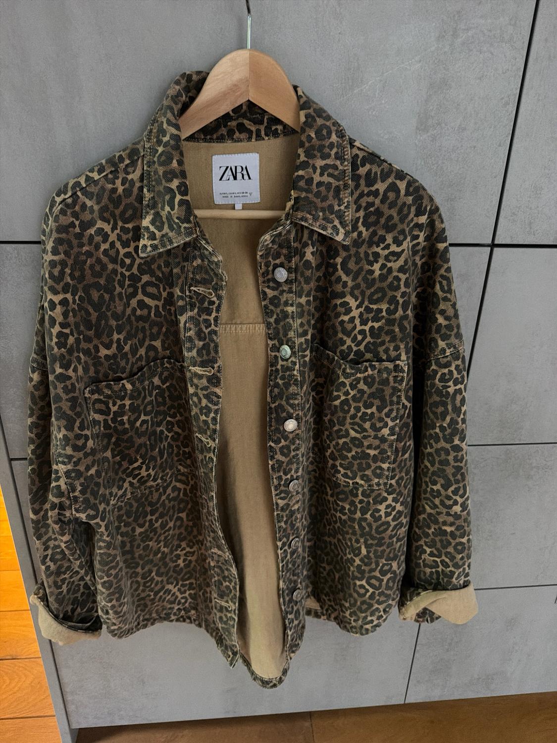 Leopard-print jacket hanging on a wooden coat hanger against a grey wall. Inner label with the text ZARA.