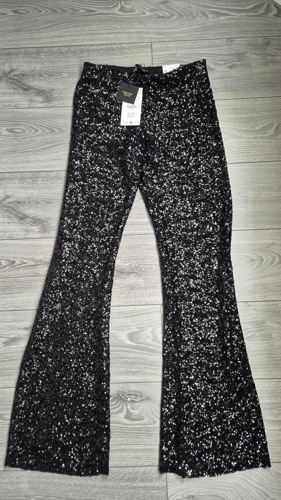 Black sequinned flared trousers on a grey wooden floor. Visible label: "FESTIVE DAYS".