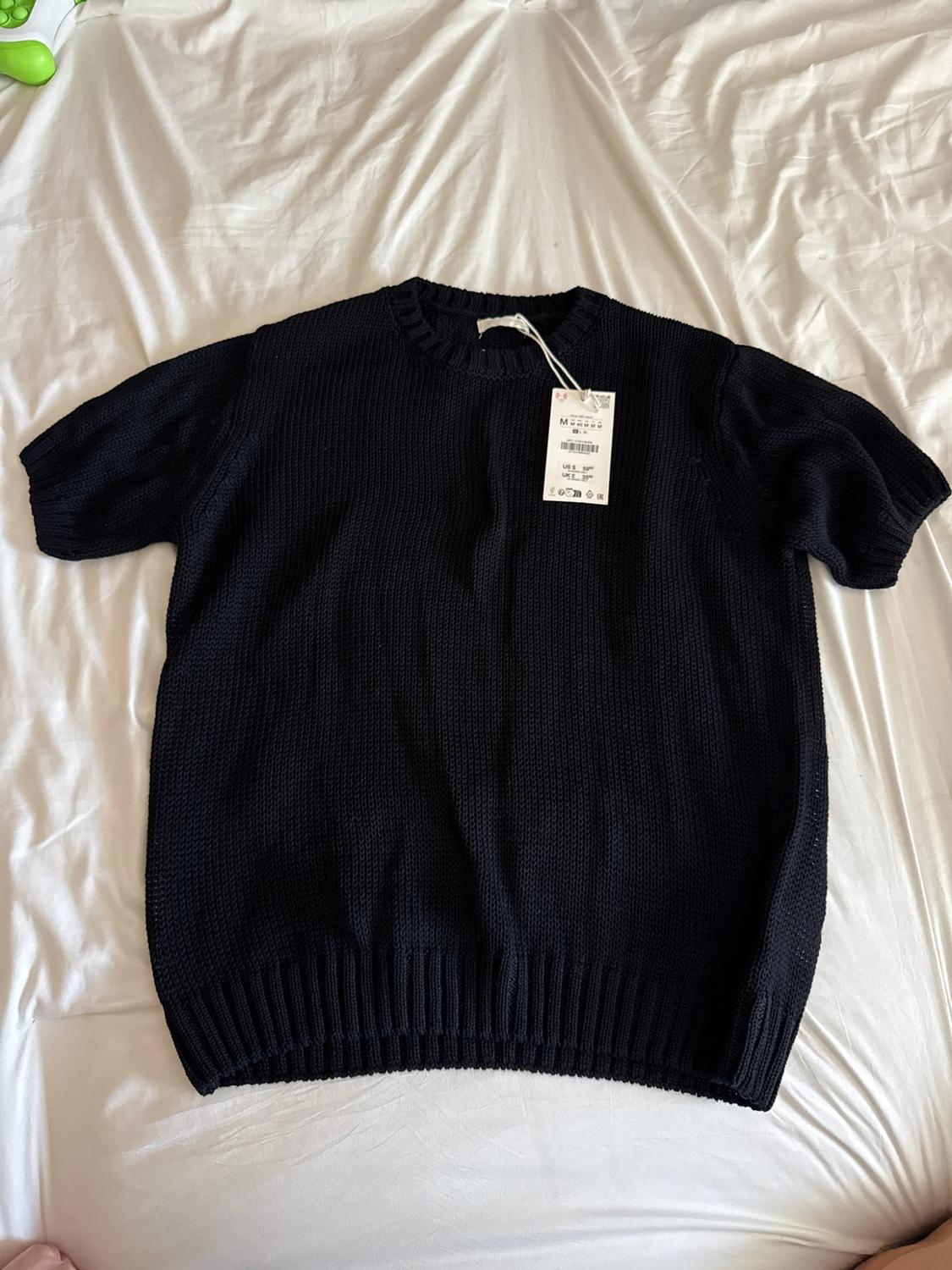 Black short-sleeve knit sweater with ribbed trim and a tag, laid out on a wrinkled white sheet.
