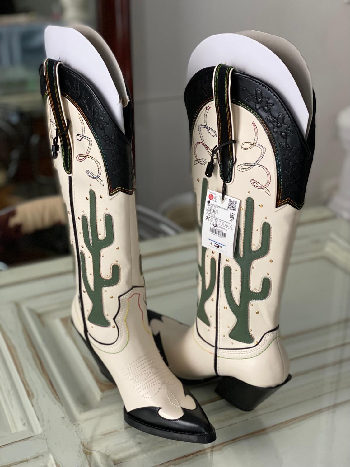 White cowboy boots with green cactus details and a black toe on a table. Visible tag with price of €89.