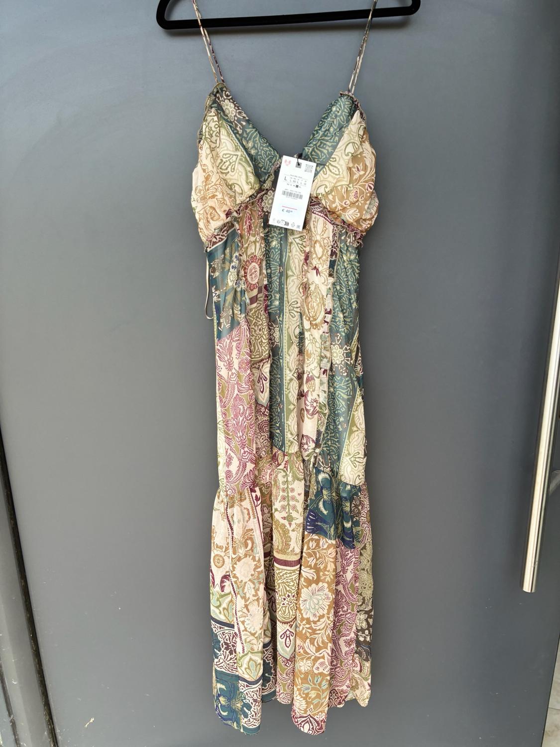 Long dress with thin straps and a floral print in shades of green, beige and burgundy, hanging on a black hanger against a grey background.