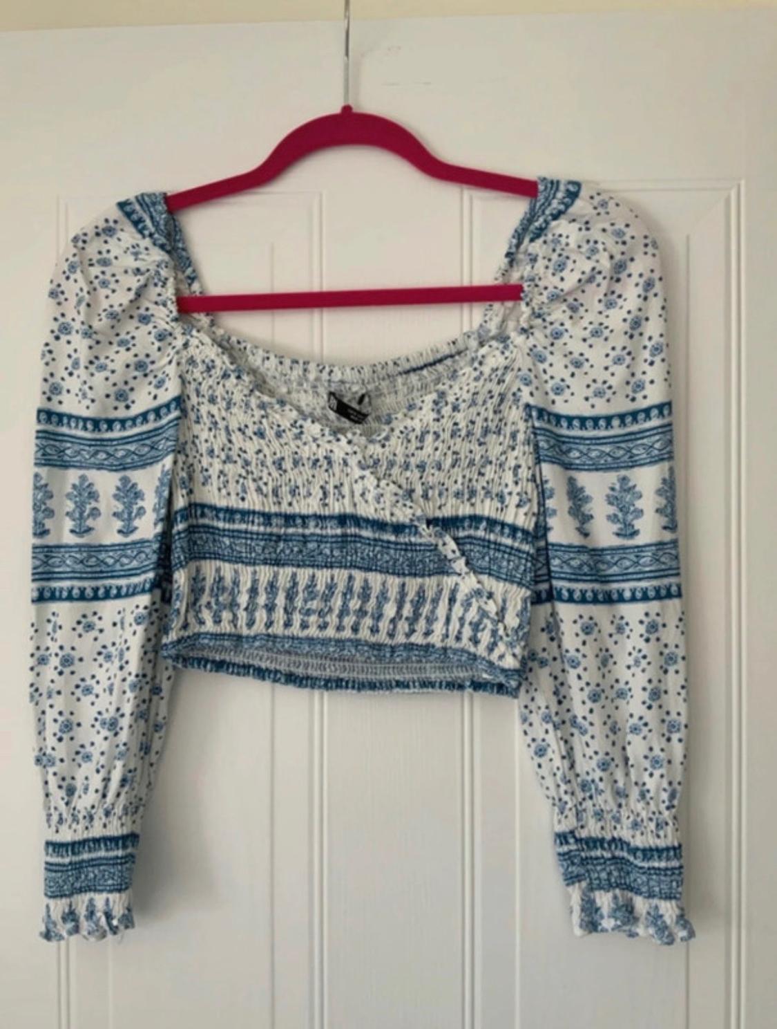 White crop top with a blue floral print, shirred bodice and puff sleeves, hanging on a pink hanger on a white door.