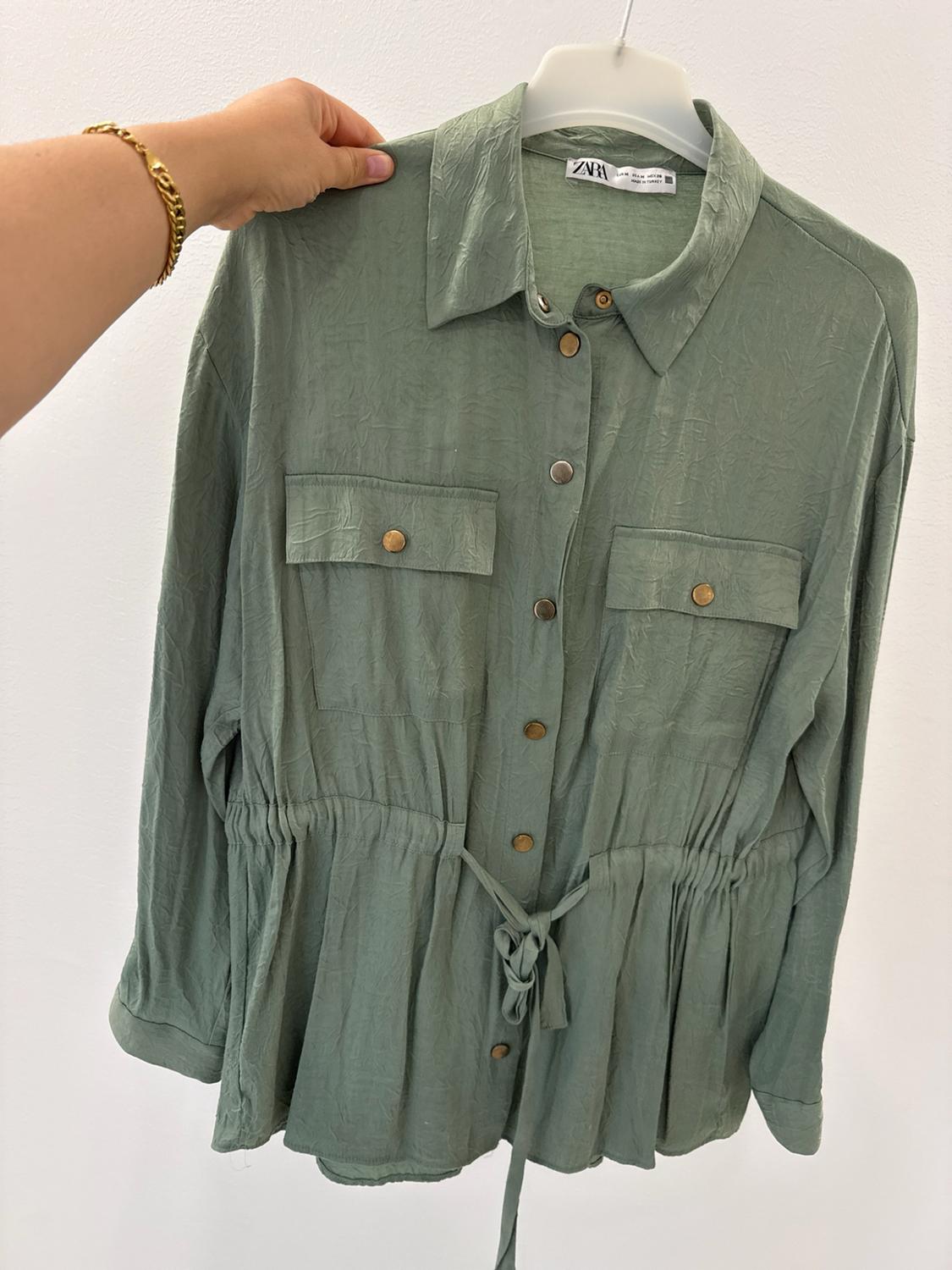 Hand holding a creased green shirt on a hanger. It has long sleeves, pockets, buttons and a drawstring waist. ZARA text.