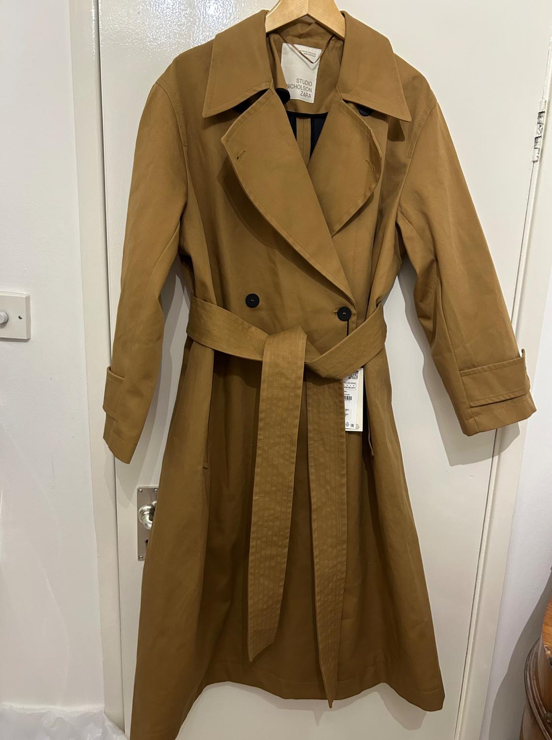Light brown trench coat with a belt and black buttons, hanging on a hanger over a white door. Label visible inside.