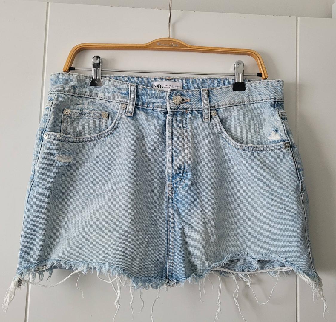Light blue denim skirt with distressed details and frayed hem, hanging on a wooden hanger.