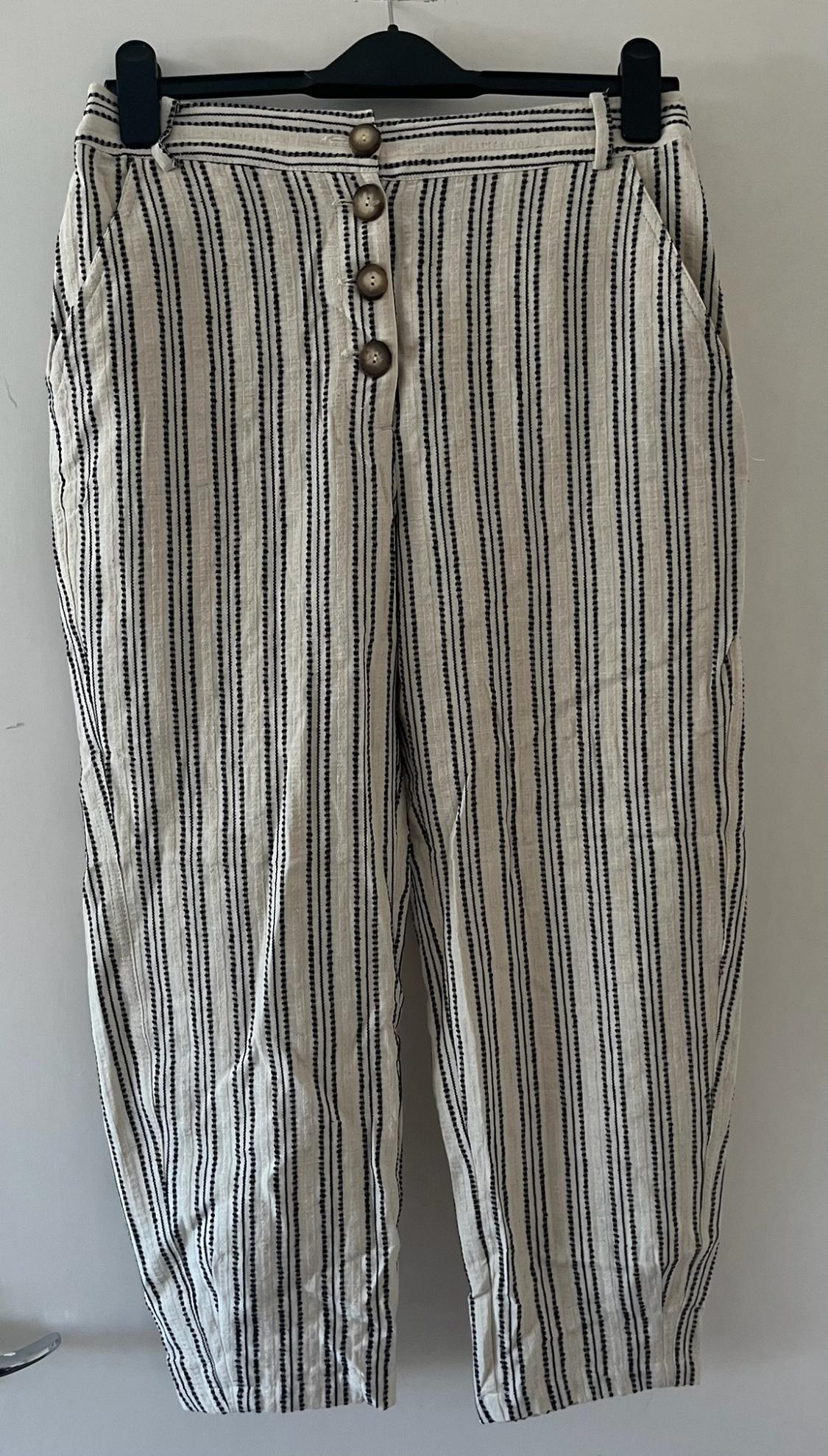 Beige trousers with vertical black stripes hanging on a black hanger against a white background. Four brown buttons stand out on the front.