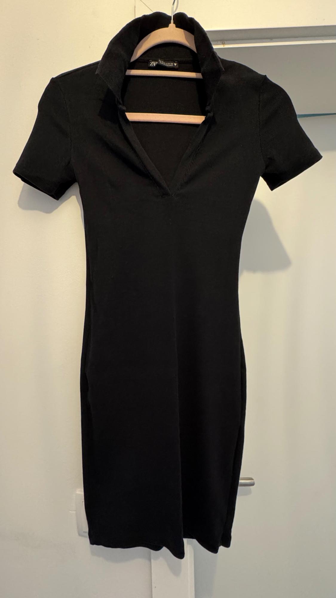 Short-sleeved black dress with an open collar, hanging on a wooden hanger against a white wall.