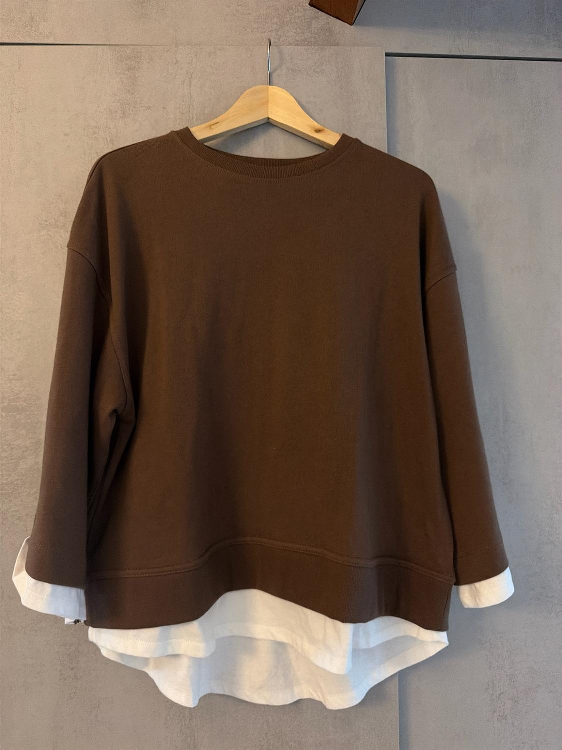 Brown sweatshirt layered over a white shirt, hanging on a wooden hanger against a grey background. Casual and relaxed style.
