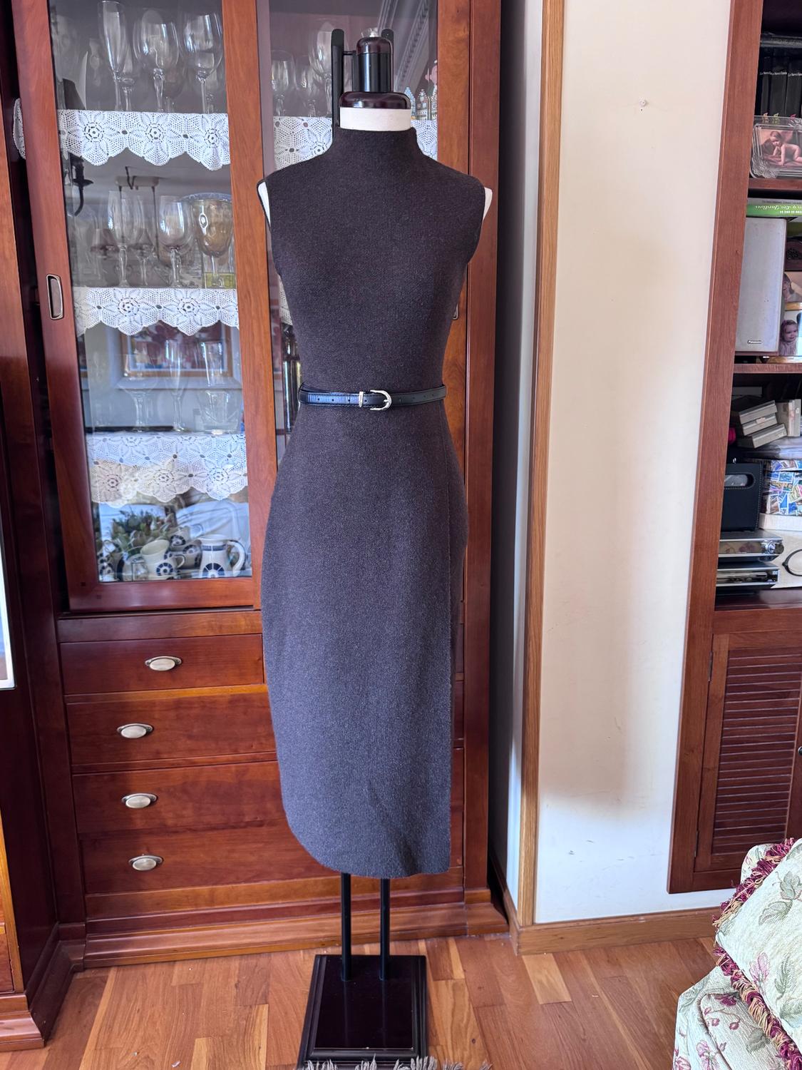 Mannequin in a grey sleeveless dress with a black belt, in front of a wooden cabinet with crystal glasses and decorative lace.