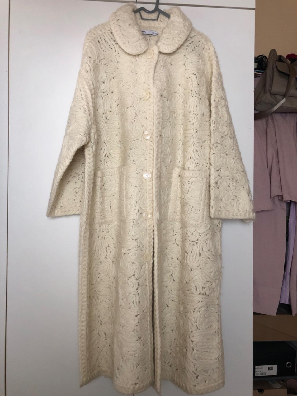 Front view of a cream long coat with crochet pattern, front buttons and side pockets.