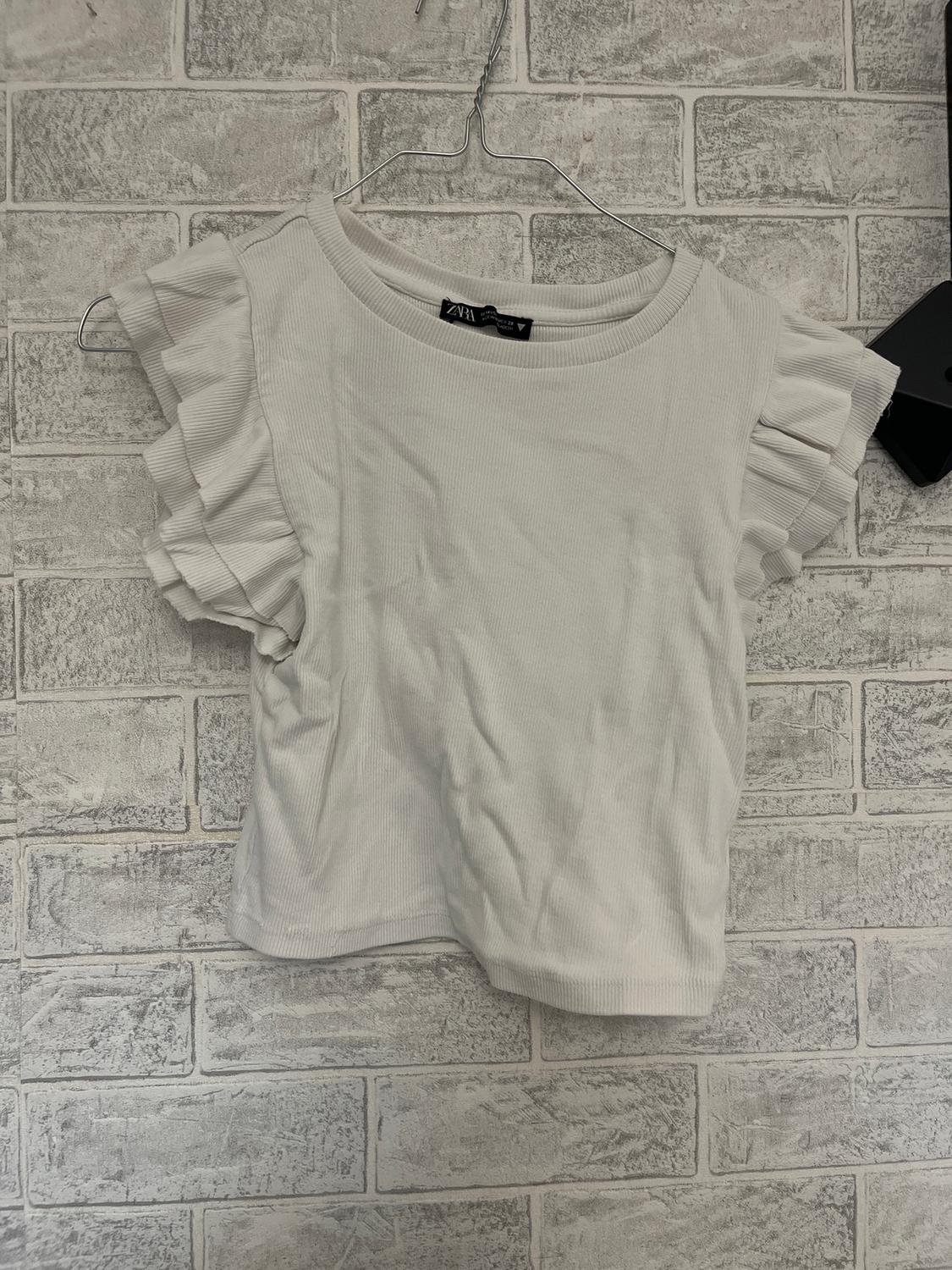 White T-shirt with ruffled sleeves, hanging on a wire hanger against a grey brick wall. Visible tag: ZARA.