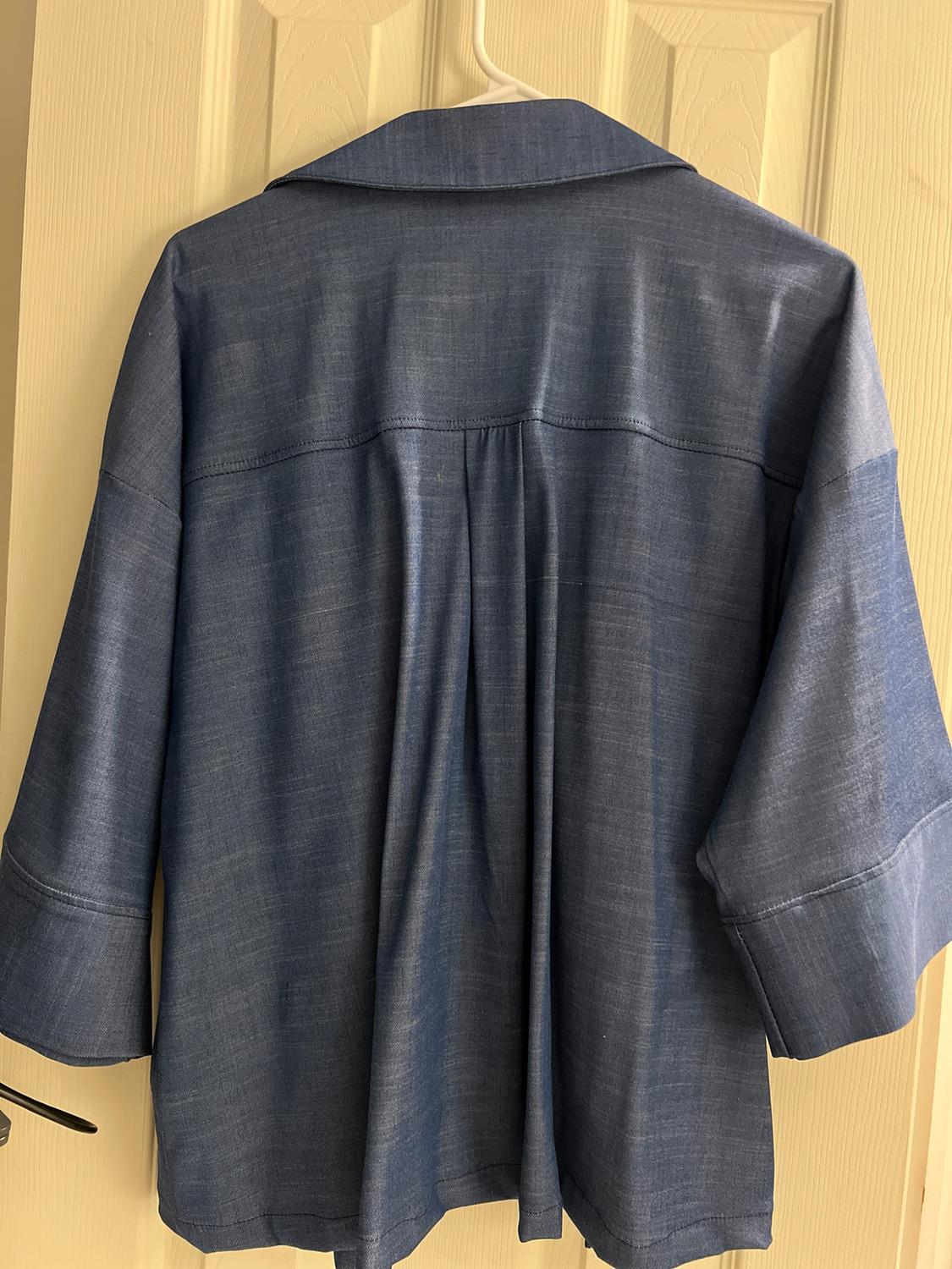 Back view of a blue shirt hanging on a white door. Its loose-fitting design with a yoke, center pleat, and wide sleeves stands out.