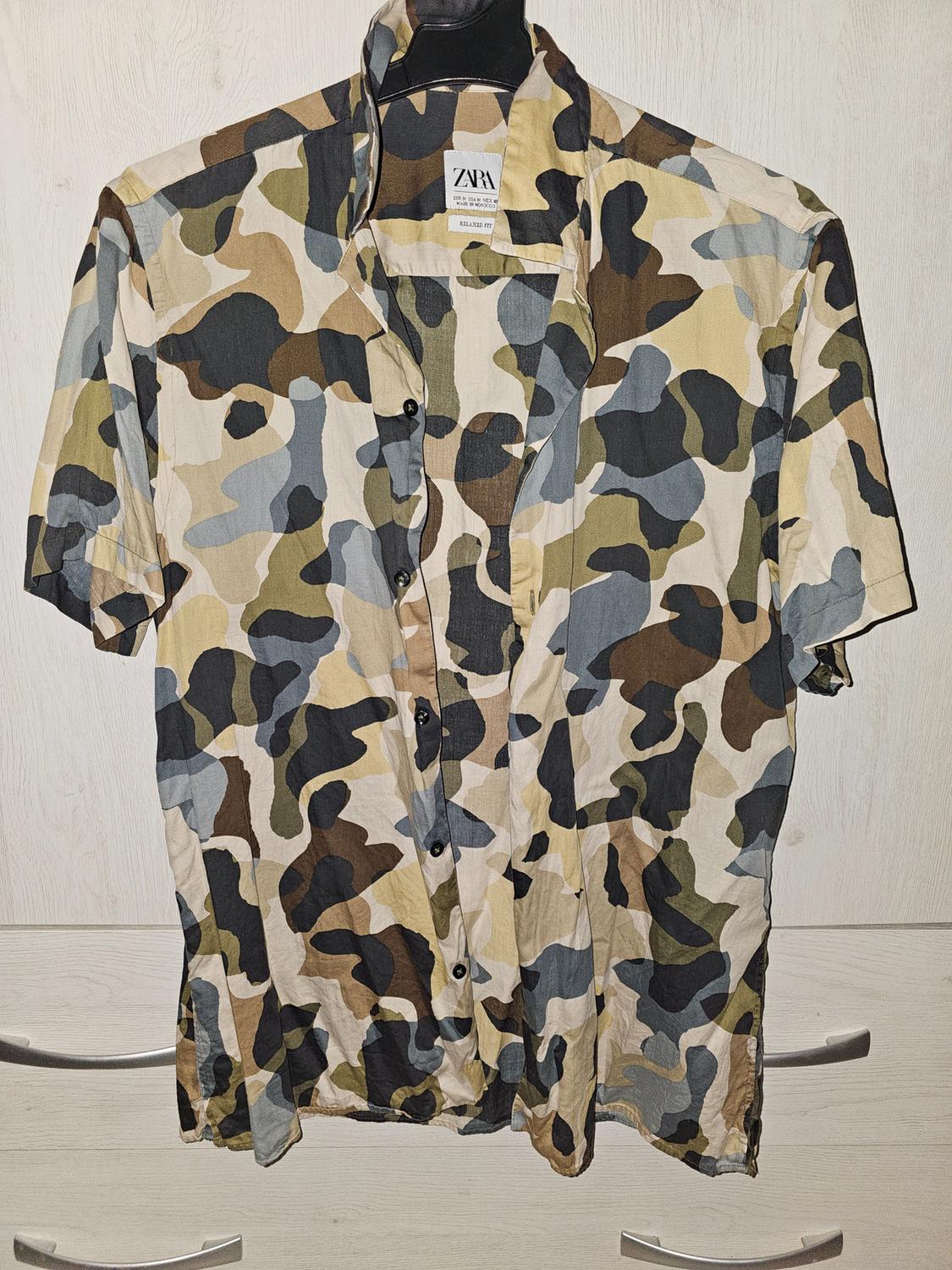 Short-sleeved shirt with a camouflage print in brown, green, and grey tones, hanging on a black hanger. Visible label: ZARA.