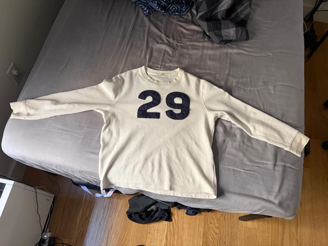 Cream long-sleeve T-shirt in color cream with the number 29 on the chest, spread out on a bed with gray sheets and a wooden floor.