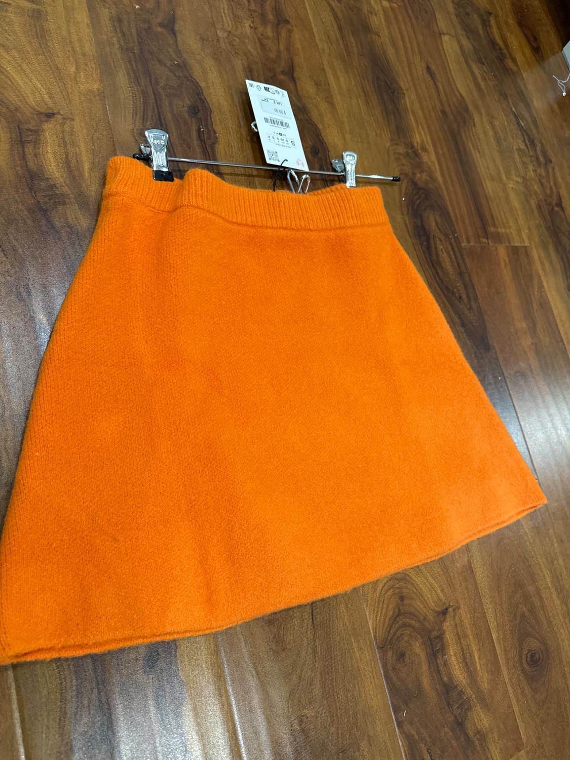 Orange mini skirt hanging on a hanger over a wooden floor. Visible label with barcode and price.