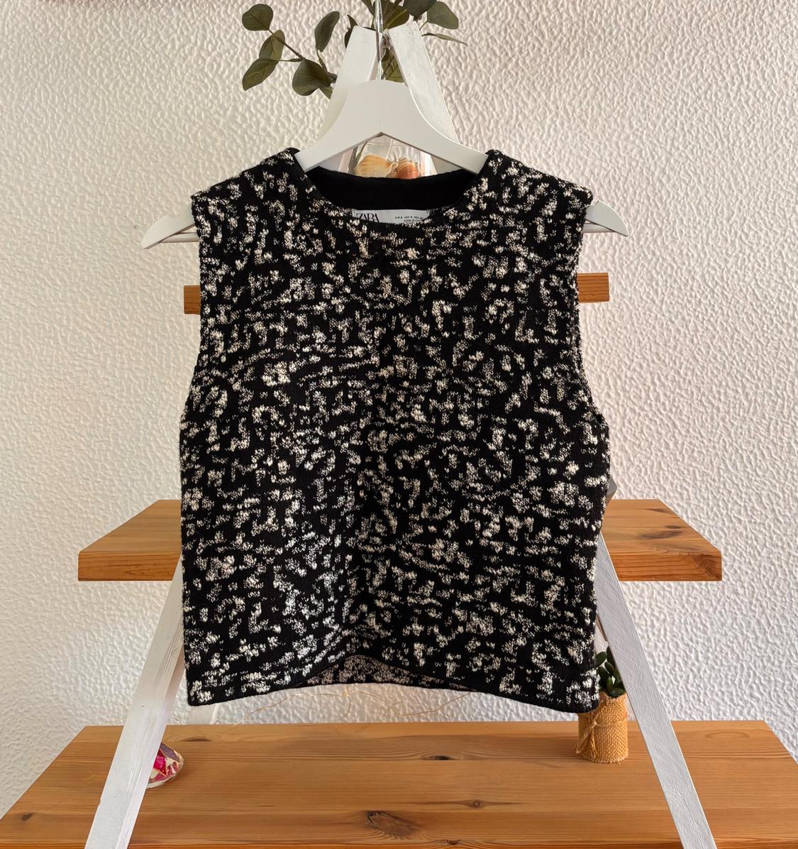 Black gilet with white print hanging on a white hanger against a textured wall background and wooden shelving.