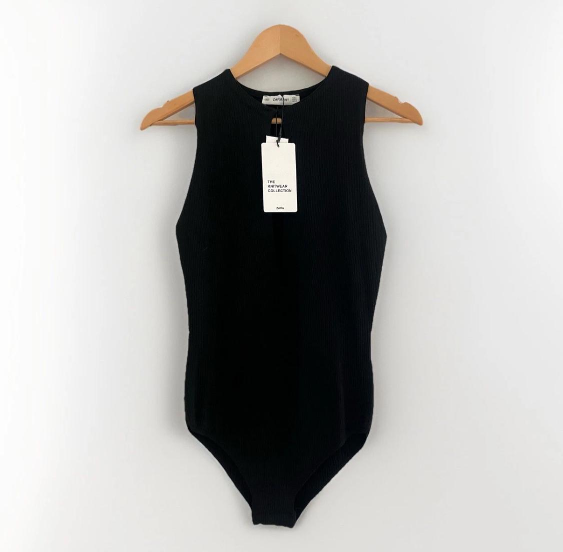 Black sleeveless bodysuit hanging on a wooden hanger against a white background. Text: ZARA THE KNITWEAR COLLECTION ZARA.