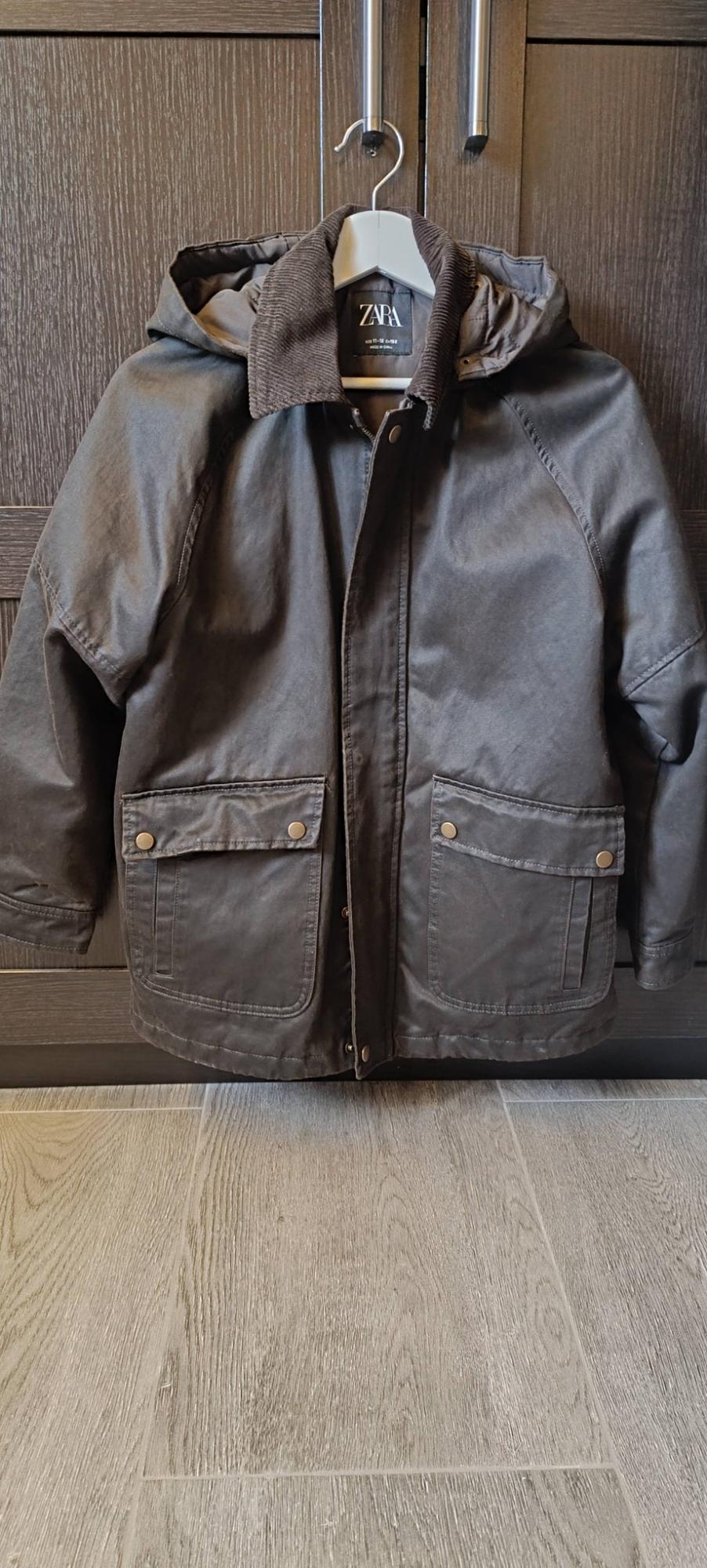 Brown hooded jacket with pockets and a corduroy collar, hanging on a white hanger in front of a wooden wardrobe. ZARA label visible.