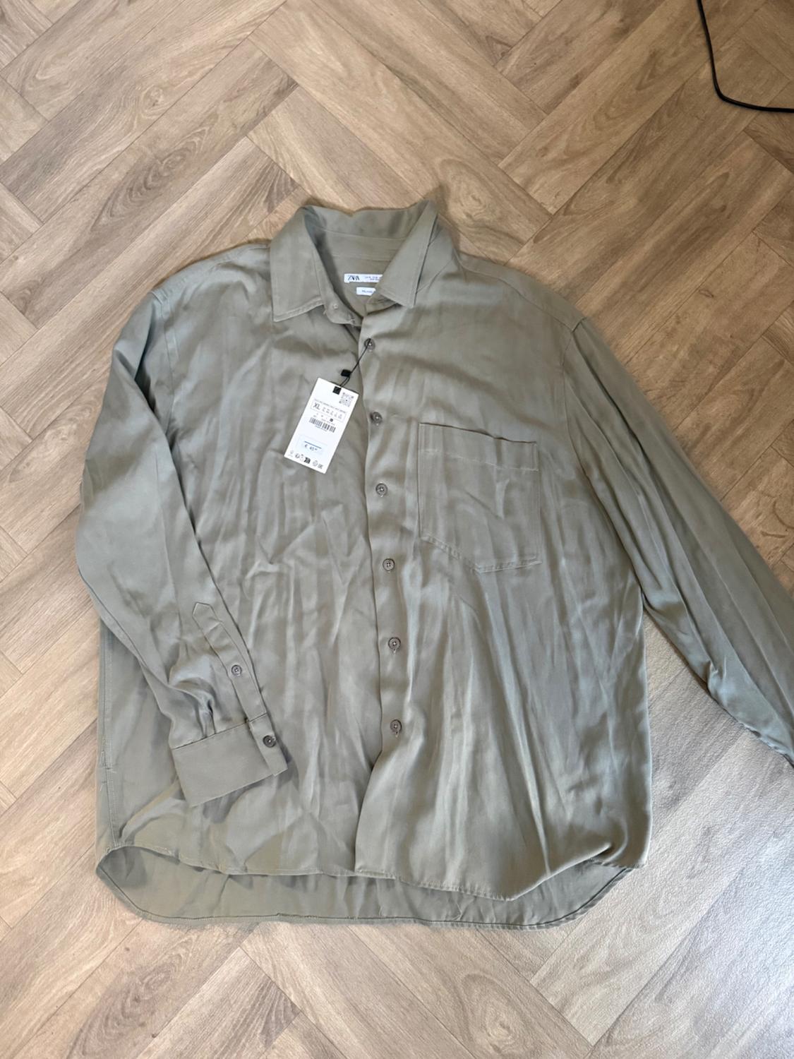 Olive green long-sleeved button-up shirt with pocket, on a herringbone wooden floor. Label with the text ZARA.