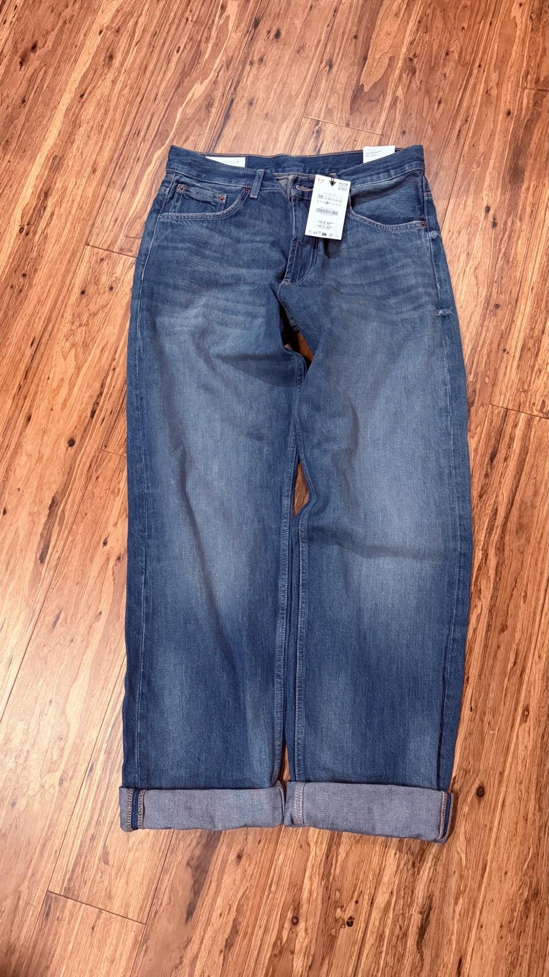 Blue denim jeans with a washed effect, cuffed hem, and a waistband label, laid flat on a wooden floor.