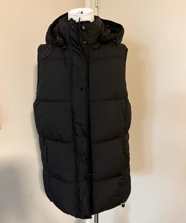 Black padded gilet with hood on a mannequin, beige background. Sleeveless design and front button fastening.