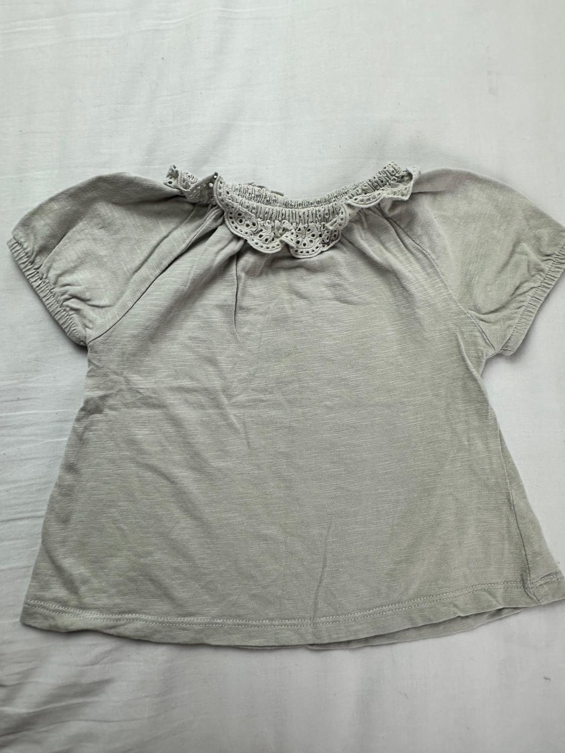 Gray short-sleeve baby T-shirt with a round neckline decorated with an embroidered lace ruffle, on a white fabric background.