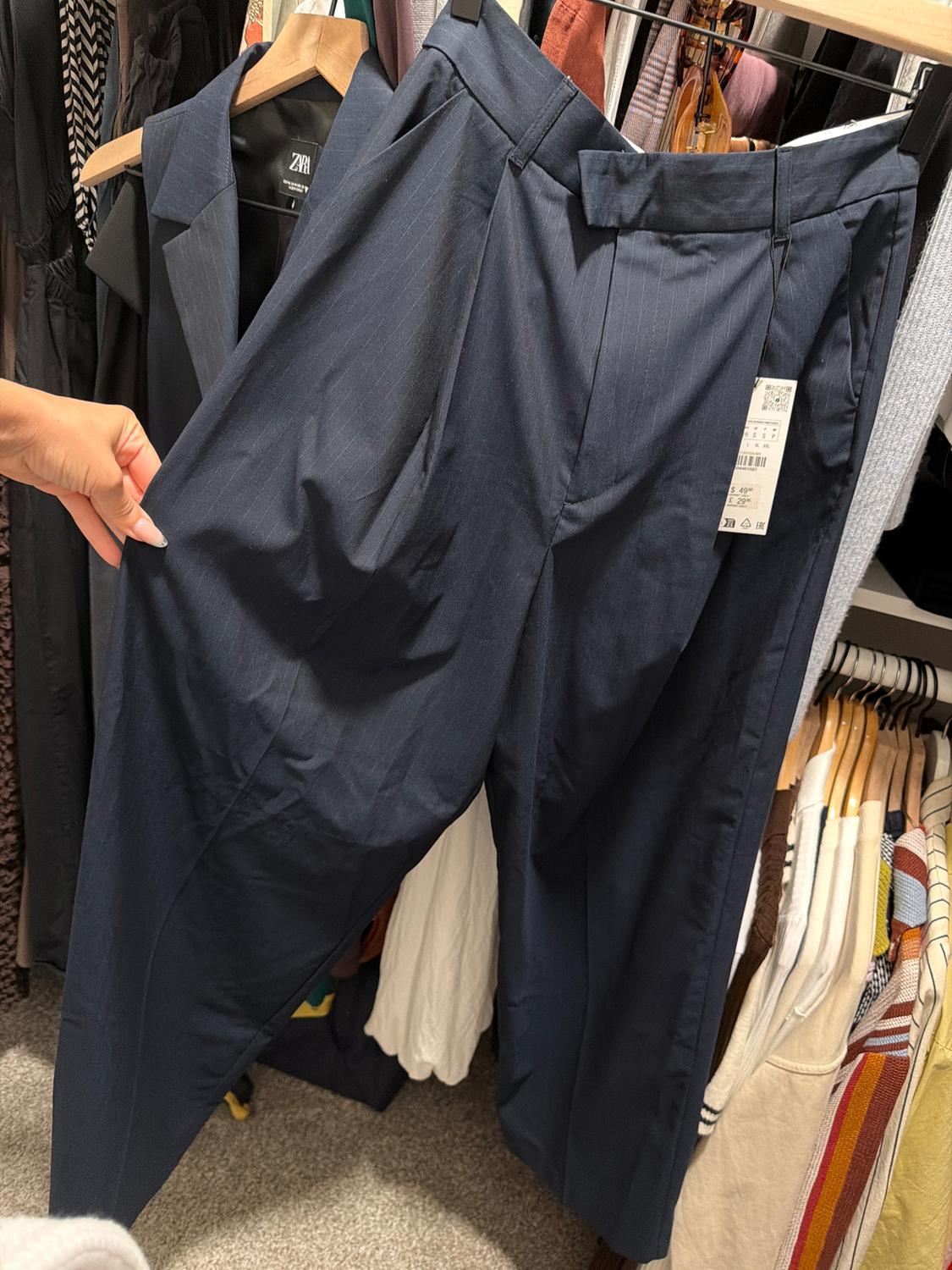 Dark blue trousers with pleats are hanging on a hanger, held by a hand. The tag is visible. The background shows various clothes.