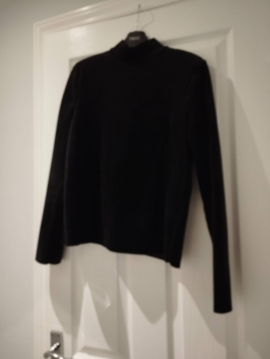 Black long-sleeved, high-neck top on a hanger, hanging from a white door. Casual style and relaxed atmosphere.