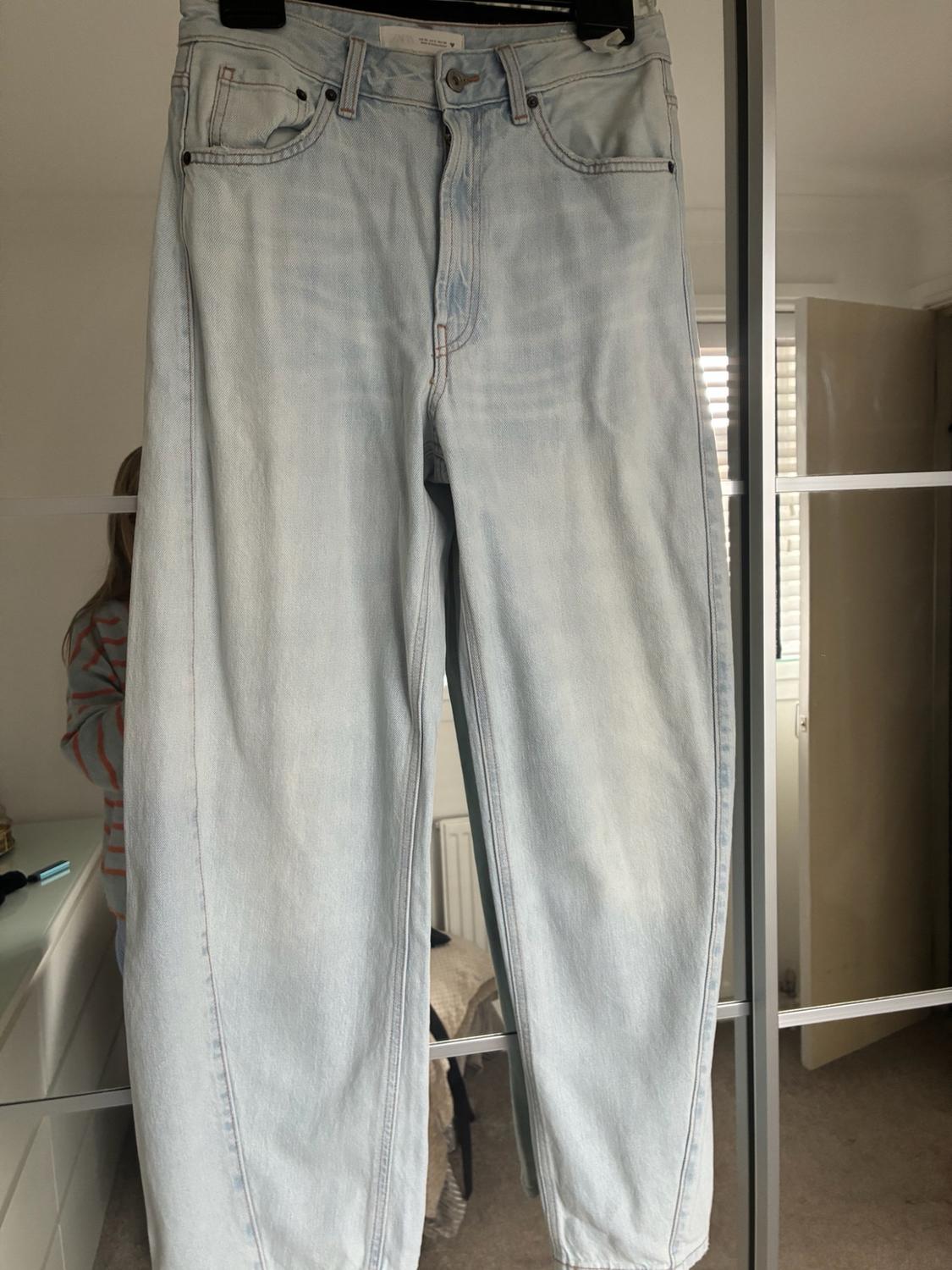 Light blue denim trousers hanging on a black hanger in front of a mirrored wardrobe reflecting part of a room and a person.