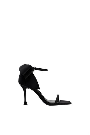 Black high-heeled shoe with an ankle strap and decorative bow on the heel, on a white background.