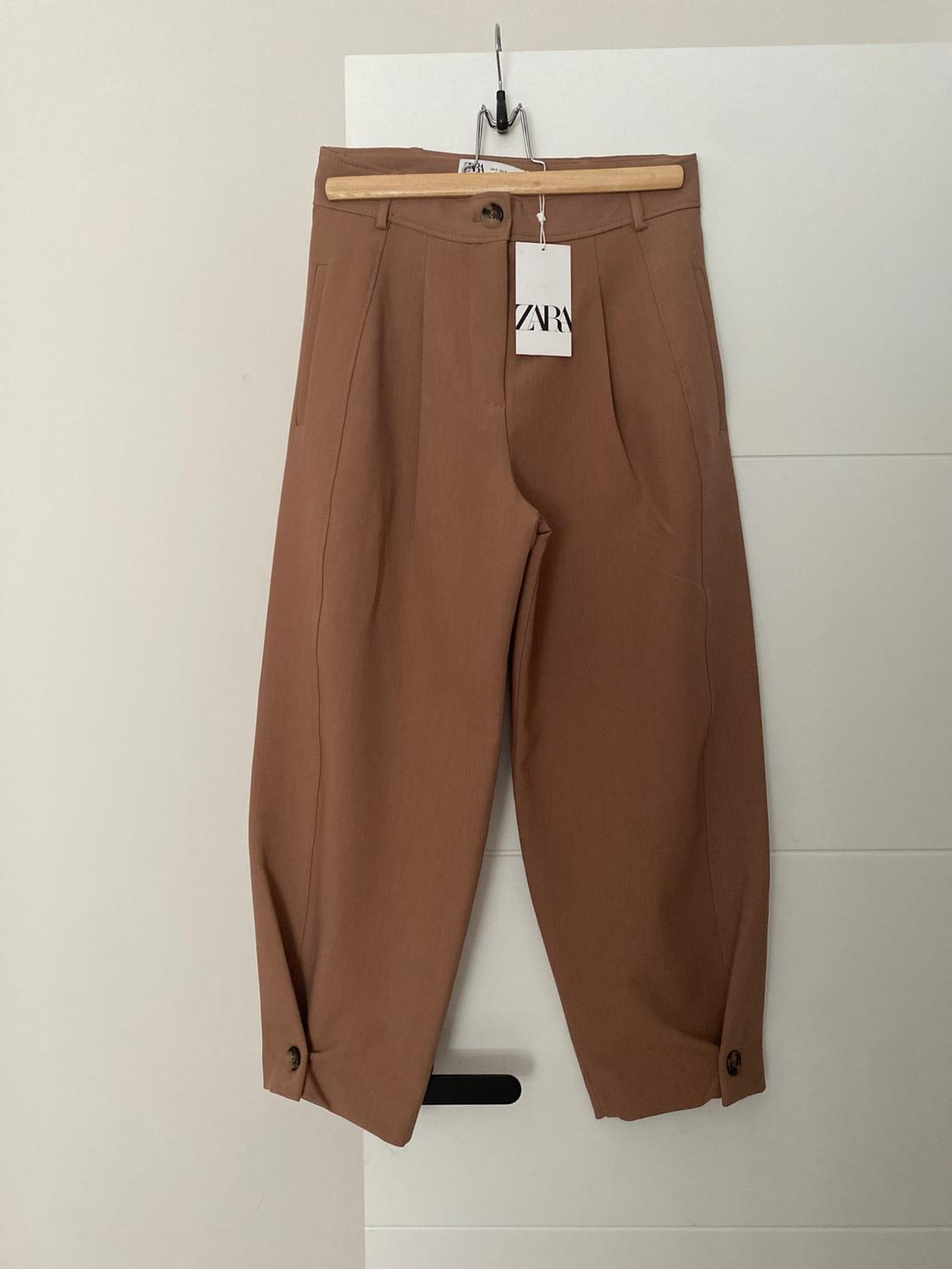 Brown tailored trousers with pleats and buttons on the hem, hanging on a wooden hanger on a white door. Label with the text ZARA.
