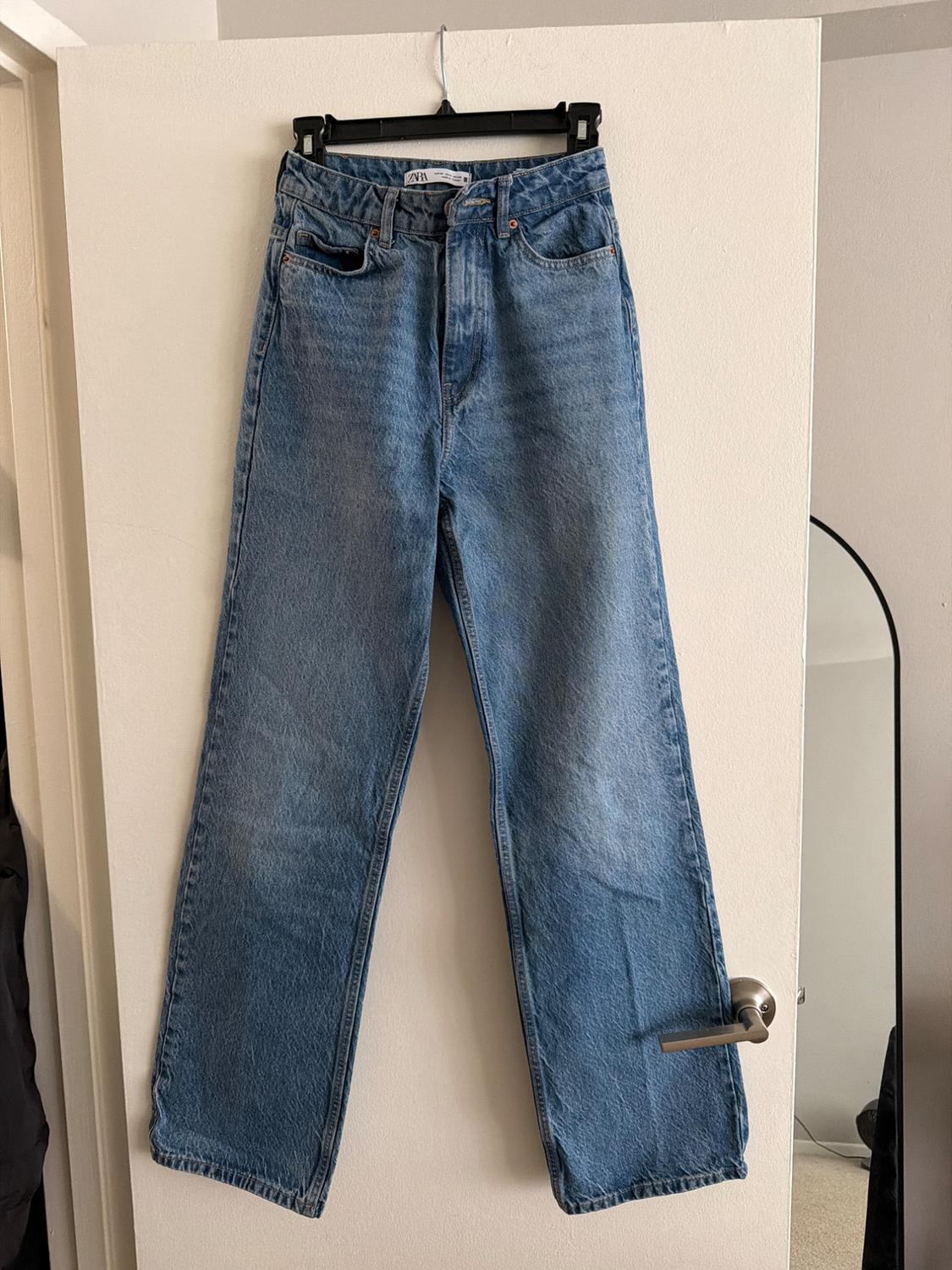 Blue jeans hanging on a black hanger over a white door. The tag shows the "Zara" brand.